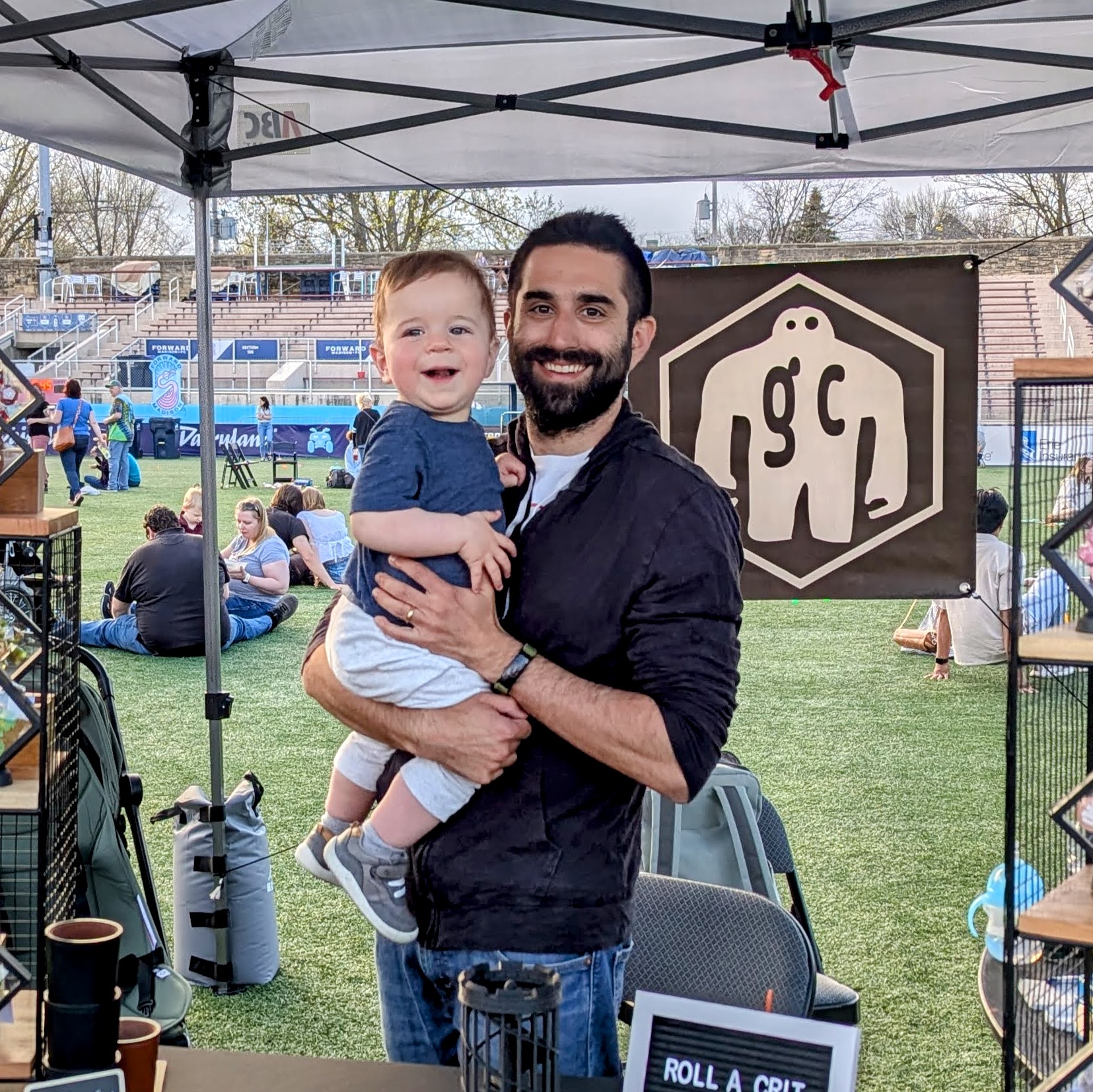 Ross at the Golem Craftworks market booth, holding a smiling toddler.